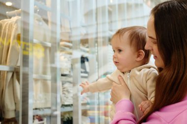 Bir anne, bir mağaza vitrininde sergilenen renkli elbiseleri heyecanlı bir şekilde işaret eden çocuğunu kucağına alır. Alışverişin ve kaynaşmanın tadını çıkarıyorlar..