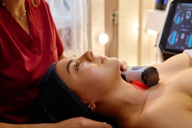 Bir kadın cilt bakımı görüyor, tedavi yatağında dinleniyor. Bir terapist, rahatlatıcı ışıklandırma ve spa dekoru ile çevrili yüz gençleştirme için bir cihaz kullanır..