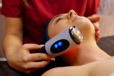Bir spa uzmanı müşteriye yüksek teknolojili yüz tedavisi uygular. Müşteri bir tedavi masasında uzanıyor, rahatlatıcı deneyimin tadını çıkarıyor ve huzurlu bir ortamda dinleniyor..