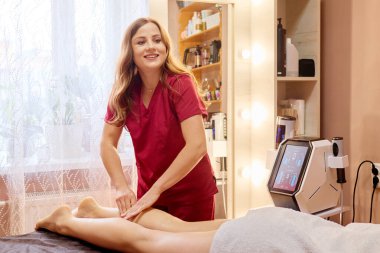 Kırmızı önlüklü bir terapist, cilt bakım ürünleriyle dolu bir stüdyoda masaj masasında yatan bir müşteri için rahatlatıcı bir bacak tedavisi uyguluyor..