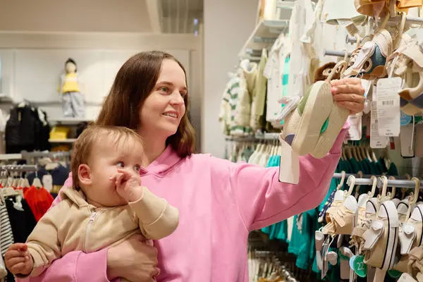 Bir anne, bebeğini sıcak bir mağazada tutarken gülümser. Çocuğu için doğru kıyafeti bulmaya çalışan bir çift sandaleti inceliyor..