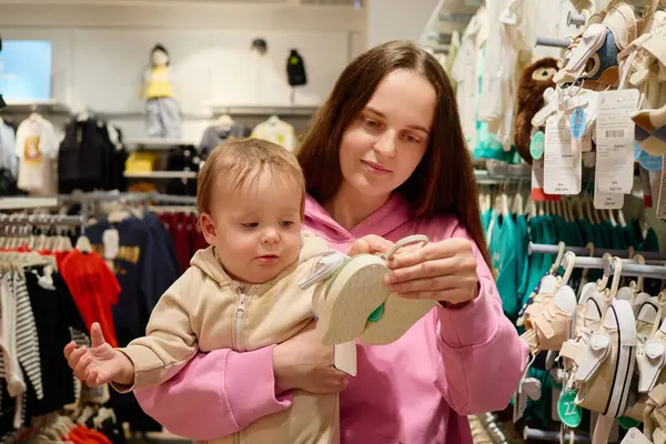 Bir anne, çocuk kıyafetleri ve aksesuarlarıyla dolu parlak, renkli bir mağazada bebek ayakkabılarını incelerken bebeğini kucağına alır. Atmosfer neşeli ve canlı..