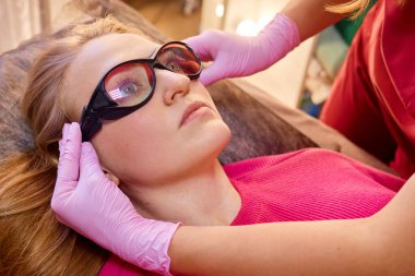 Bir müşteri salonda bir tedavi yatağında uzanıyor, deri ameliyatına hazırlanıyor. Eldiven giymiş bir pratisyen, müşterinin yüzündeki koruyucu gözlüğü dikkatlice ayarlar..