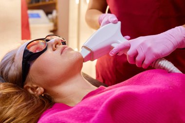 Çağdaş bir güzellik salonunda, bir kadın koruyucu gözlük takarak bir tedavi yatağında yatar. Bir güzellik uzmanı, çenesinde cilt bakımı için bir cihaz kullanırken aynı zamanda gevşek ifadesini de vurguluyor..