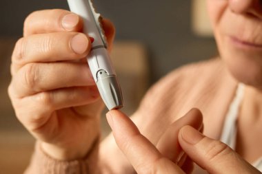 Yaşlı bir birey parmak ucuna yakın bir kan şekeri testi kalemi tutarak glikoz seviyelerini kontrol etmeye hazırlanıyor. İçerideki sıcak ortam rutin bir sağlık kontrolüne işaret ediyor..