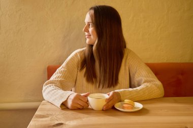 Uzun saçlı bir kadın sıcak bir kafede oturuyor, bir elinde bir fincan çay tutuyor ve dikkatlice pencereden dışarı bakıyor. Masanın yanında renkli bir tabak pasta var..