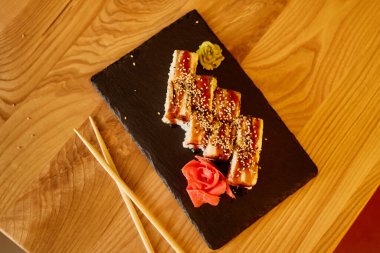 Sushi dürümleri siyah bir tabakta, susam tohumlarıyla kaplı ve yanında bir gül süslemesi ile güzelce düzenlenir. Taze wasabi sunuma canlı bir dokunuş ekler..