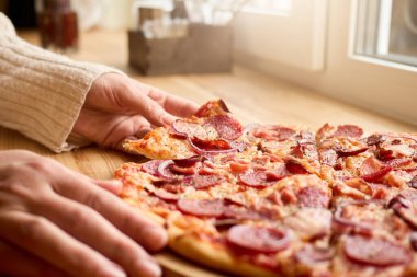 Biri ahşap bir masaya yerleştirilmiş pepperoni pizzasına uzanır. Dekor yumuşak, doğal bir ışık içeriyor, rahat restoranın davetkar havasını arttırıyor..