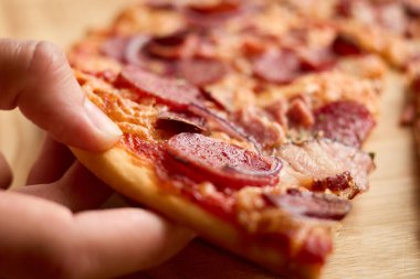 Bir dilim pepperonili pizzaya uzanan bir elin yakınına. Altın hamuru eritilmiş peynir, baharatlı pepperoni ve otlarla kaplı..