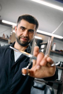 Yetenekli bir berber gülümseyerek makas tutar. Süslü bakım stüdyosunda, çeşitli saç ürünleriyle dolu raflar var. Parlak tepelik ışıklarla aydınlatılmış..