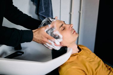 Bir erkek kuaförde rahatlatıcı bir şampuan tedavisinden hoşlanır. Bir stilist nazikçe kafa derisine masaj yapar kuaför lavabosundan su akarken, öğleden sonra ışığında sakin bir deneyim yaratır..