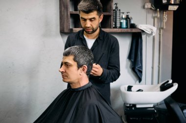 Bir berber, bir müşterinin saçını modern bir kuaför ortamında titizlikle şekillendirir. Berber işine odaklanırken müşteri sabırla pelerin giyer ve etrafı tımar ürünleriyle çevrilidir..