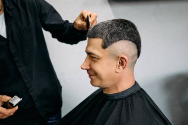 Bir berber modern bir berber dükkanında bir adamın saçını titizlikle şekillendirir. Adam siyah bir pelerin içinde oturuyor, berber keskin bir bakış oluşturmaya odaklanırken gülümsüyor..