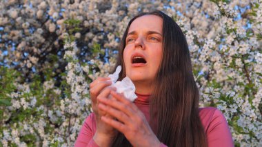 Çiçek açan ağaçların arasında duran bir kadın, bahar boyunca elinde bir mendil tutarken polen alerjilerinden görünür bir şekilde etkilenir..