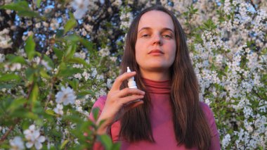 Kadın bahar çiçeklerinin arasında duruyor, bahçedeki polenlerin yol açtığı semptomları hafifletmek için alerji spreyi kullanıyor..