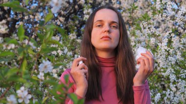 Bahar bahçesinde beyaz çiçeklerin arasında duran bir kadın, her iki elinde de dokular olan polen alerjilerinden etkilenir..