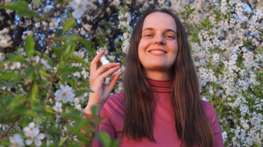 Bir kadın güzel bahar çiçeklerinin arasında duruyor, polen alerjisine değinirken gülümsüyor güneşli bir bahçede özenle.