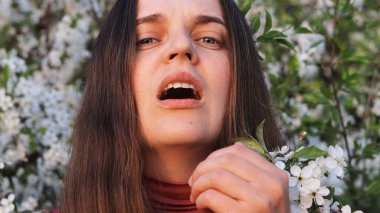 Çiçeklerin arasında duran bir insan, alerjik reaksiyonlar sergiliyor. Yüzünde, doğa tarafından sarılmış, gözle görülür bir rahatsızlıkla..