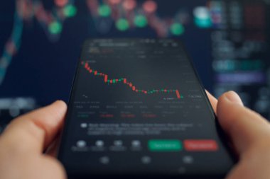 Bir kişinin elinde azalan bir borsa grafiğini ağırlıklı olarak kırmızı mumlarla gösteren, aşağı yönlü bir eğilim gösteren, daha büyük ekranda daha fazla finansal veriyi bulanık gösteren, daha büyük ekranda mobil ticareti öneren bir akıllı telefon var. 