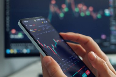 Bir kişinin elleri, daha büyük bir ekranda daha fazla finansal veri gösteren, mobil ticaret veya yatırım analizine işaret eden, kırmızı ve yeşil mumları olan, fiyat dalgalanmalarını gösteren bir akıllı telefon tutuyor.