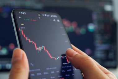 Bir kişinin elleri, daha büyük bir finansal veri ekranının bulanık zeminine karşı çoğunlukla kırmızı şamdanlarla azalan bir borsa grafiğini gösteren ve pazar düşüşü sırasında mobil ticareti gösteren bir akıllı telefon tutuyor.