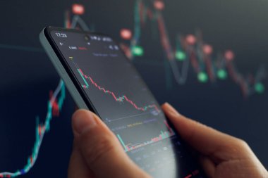 Bir el, benzer verilerin bulanık arkaplanına karşı azalan borsa grafiğini gösteren bir akıllı telefonu tutuyor.