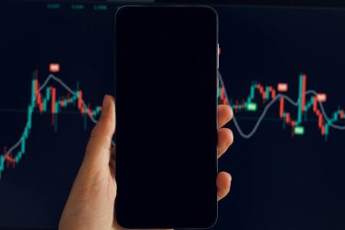 Bir el, kırmızı ve yeşil şamdanlarla dalgalanan bir borsa tablosunu gösteren akıllı telefonu tutuyor. Başka bir bulanık ekran arka planda benzer verileri gösterir.