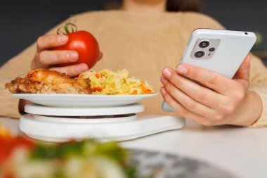 Domatesleri mutfak terazisinde tartarken akıllı telefon kullanmak. Sağlıklı beslenme, porsiyon kontrolü ve diyet izleme teknolojisi.