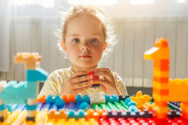 Parlak gözlü genç kız renkli oyuncak setinden bir blok ötede kameraya bakıyor. Çocukluk, yaratıcılık ve oyun.