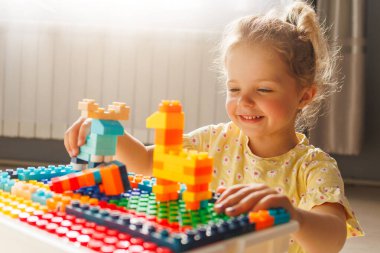 Gülümseyen genç kız parlak güneş ışığında renkli oyuncak inşaatçılarıyla mutlu bir şekilde inşa ediyor, yaratıcı oyunlardan ve inşaatlardan zevk alıyor..