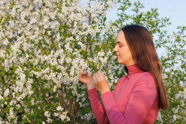 Kadın dışarıda beyaz çiçekleri huzur içinde kokluyor, sakin ve alerjisi olmayan, doğanın güzelliğinin ve taze bahar havasının tadını çıkaran..
