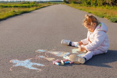Açık bir yolda renkli bir tebeşirle oturan genç kız, kırsal alanda çocukluk yaratıcılığını, eğlencesini ve geçici sokak sanatını gösteriyor..