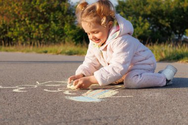 Pembe ceketli gülümseyen bir çocuk asfaltta beyaz tebeşirle resim çizer, güneşli bir günde açık havada oynamaktan zevk alır..