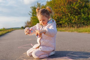 Pembe ceketli şirin bir kız asfalta oturmuş, güneşli bir günde çizimlerle çevrili renkli tebeşirleri tutmaya ve incelemeye odaklanmış..