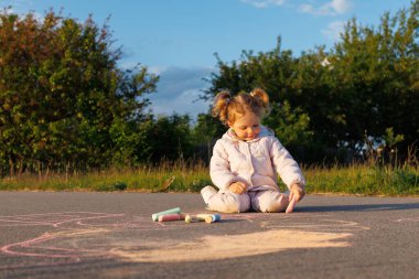 Pembe ceketli bir çocuk asfaltta oturur, renkli tebeşirle çizer, açık havada güneşli bir günde sanatına dalmıştır..