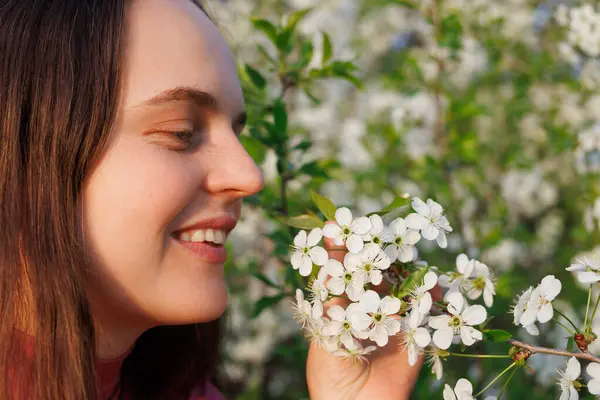 Dışarıdaki beyaz çiçekleri koklayarak gülümseyen, alerjisi olmayan, bahar aylarında doğanın kokusundan zevk alan bir kadın..