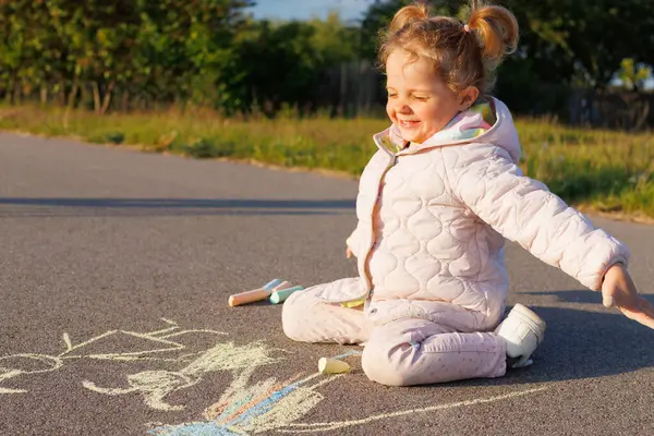 Pembe ceketli mutlu bir çocuk asfaltta oturur, renkli tebeşirle çizer, açık havada güneşli bir günün tadını çıkarır..