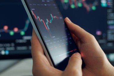 Borsa eğilimlerini ve veri analizlerini gösteren bir akıllı telefon ile etkileşime giren kişi, arka planda ek finansal grafiklerle çevrilidir..