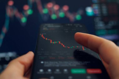 Bir kişi borsa verilerini gösteren akıllı bir telefonla etkileşime girer, yakınlardaki bir bilgisayar ekranında güncellenen grafikler gibi finansal analize odaklanır.