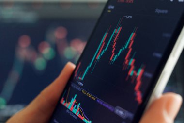 Bir kişi akıllı telefondan hisse senedi eğilimlerini analiz eder, şamdan çizelgeleri ve ticari verileri inceler ve piyasa göstergeleri arkasında bulanıklaşır.