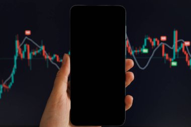 Arka planda renkli bir ticaret grafiği değişen pazar eğilimleri ve göstergeleri gösterirken, bir kişi boş bir akıllı telefon tutuyor.