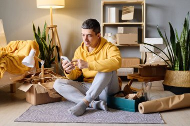 Sarı kapüşonlu bir adam desenli bir halıda bağdaş kurarak oturuyor. Akıllı telefonunu paketleri açılmamış kutular ve kapalı alandaki bitkilerin arasında kullanıyor..