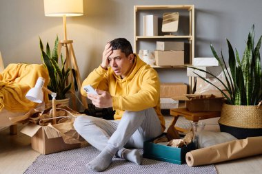 Bir adam kutuların arasında oturmuş telefonuna stresli bir şekilde bakıyordu. Yalnız yaşamanın zorlukları.