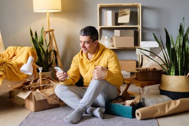 Bir adam, yeni evine taşındıktan sonra akıllı telefonuna gülümseyerek bakarak kutuları taşırken yerde oturuyor..