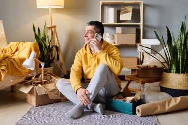 Bir adam yeni evine taşındıktan sonra kutuları taşırken yerde oturmuş telefonuyla mutlu bir şekilde konuşuyor..