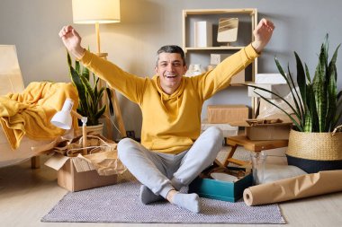 Sarı kapüşonlu neşeli bir adam kollarını kaldırmış bir halının üzerinde, kutuları ve bitkilerle çevrili bir şekilde oturuyor..
