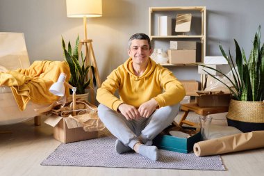 Gülümseyen bir adam, bacakları bağlı bir şekilde yerde oturuyor. Yeni dairesinde paketlenmemiş kutular arasında, mutlu görünüyor..