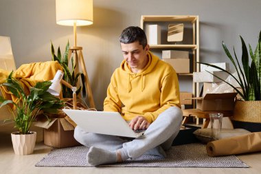 Bir adam yeni evine taşındıktan sonra dizüstü bilgisayarını kullanarak kutuları taşırken yerde oturuyor..