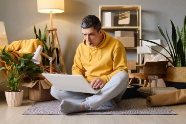 Sarı kapüşonlu, şaşkın bir adam dizüstü bilgisayarı açık kutularına ve kapalı alandaki bitkilere bakıyor..