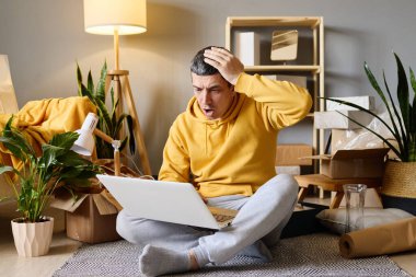 Bir adam, yeni evine taşındıktan sonra dizüstü bilgisayarına şok olmuş bir şekilde, kutuları taşırken yerde oturuyor..